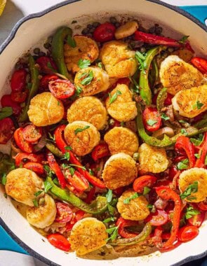 Mediterranean scallops nestled with tomatoes and bell peppers in a skillet. Next to this is a cloth napkin, a lemon, and a bowl of chopped parsley.