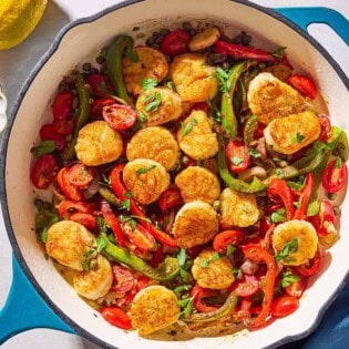 Mediterranean scallops nestled with tomatoes and bell peppers in a skillet. Next to this is a cloth napkin, a lemon, and a bowl of chopped parsley.