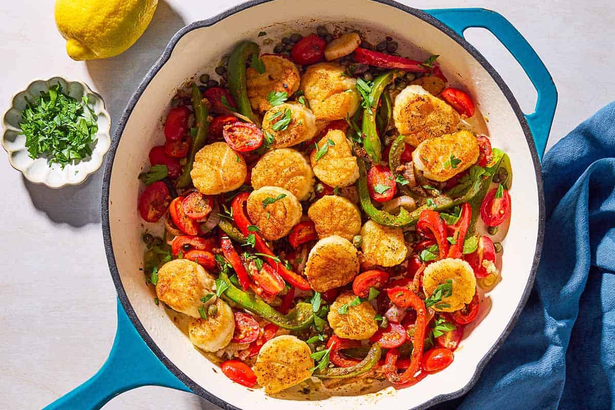 Mediterranean scallops nestled with tomatoes and bell peppers in a skillet. Next to this is a cloth napkin, a lemon, and a bowl of chopped parsley.