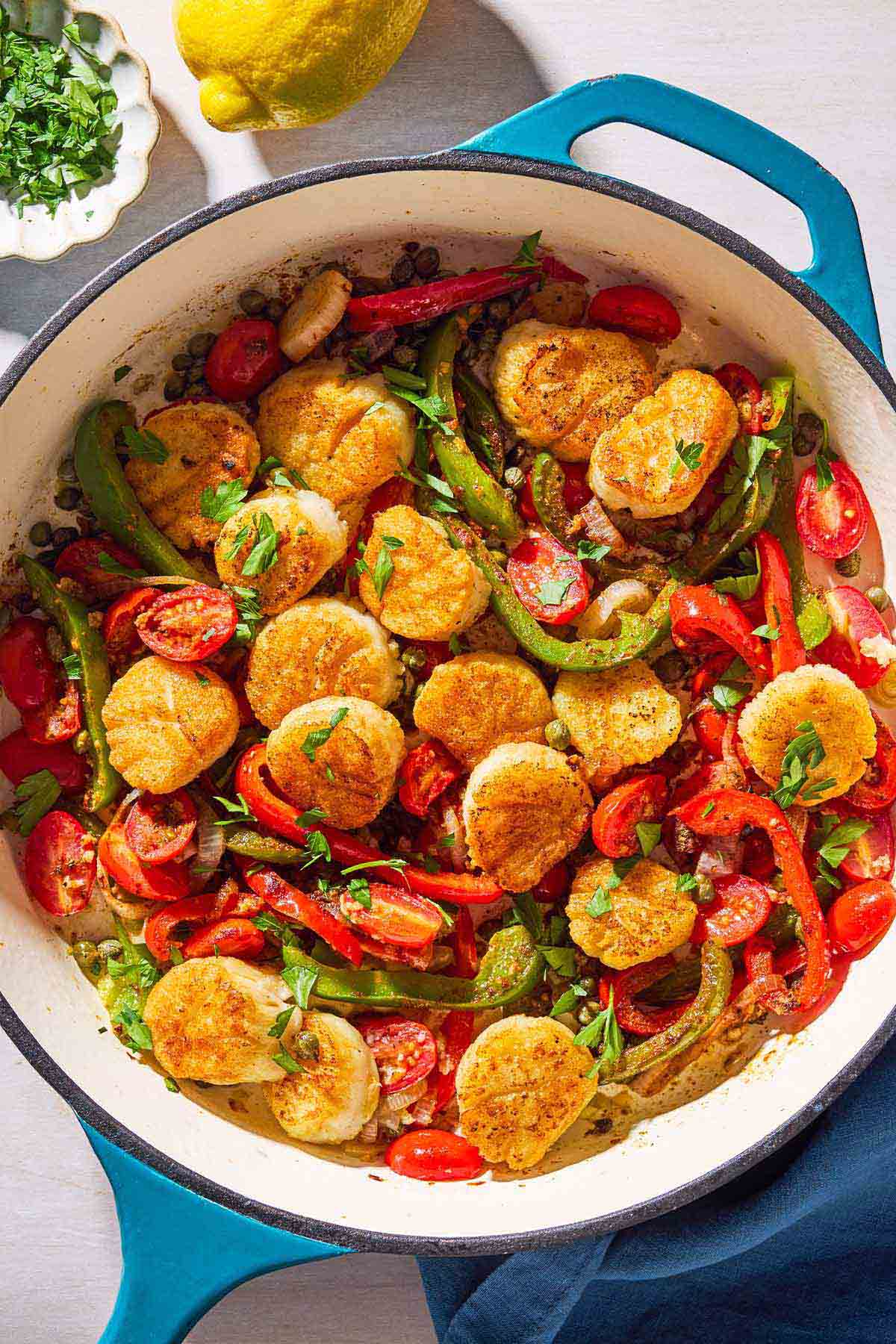 Mediterranean scallops nestled with tomatoes and bell peppers in a skillet. Next to this is a cloth napkin, a lemon, and a bowl of chopped parsley.