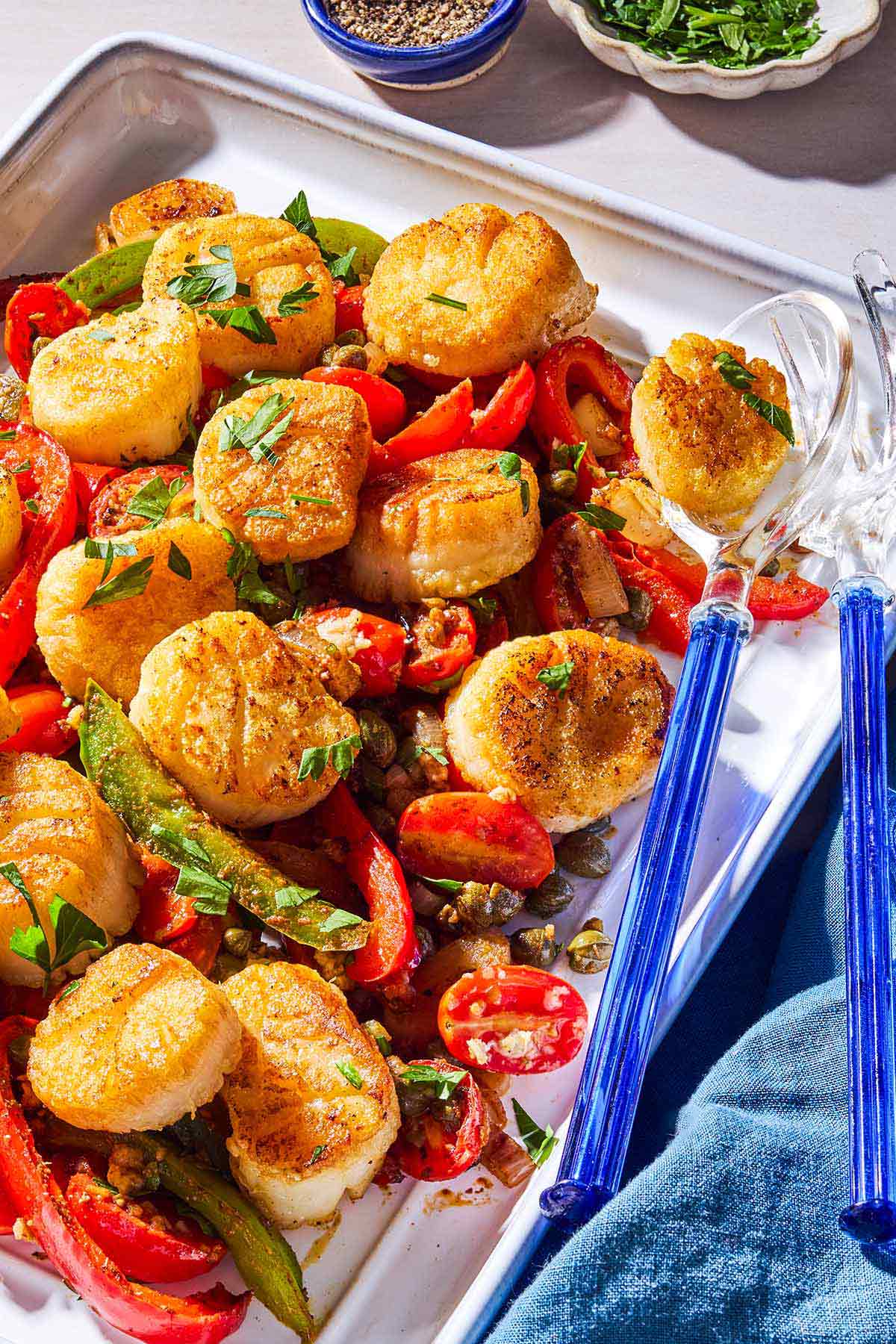 Mediterranean scallops nestled with tomatoes and bell peppers on a serving platter with serving utensils.