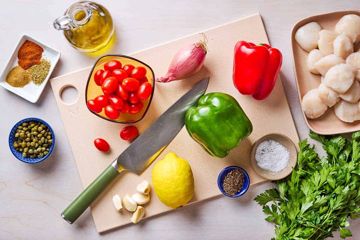 Ingredients for Mediterranean scallop recipe including scallops, olive oil, a shallot, a red pepper, a green pepper, garlic, grape tomatoes, capers, salt, black pepper, oregano, cumin, paprika, lemon and parsley.