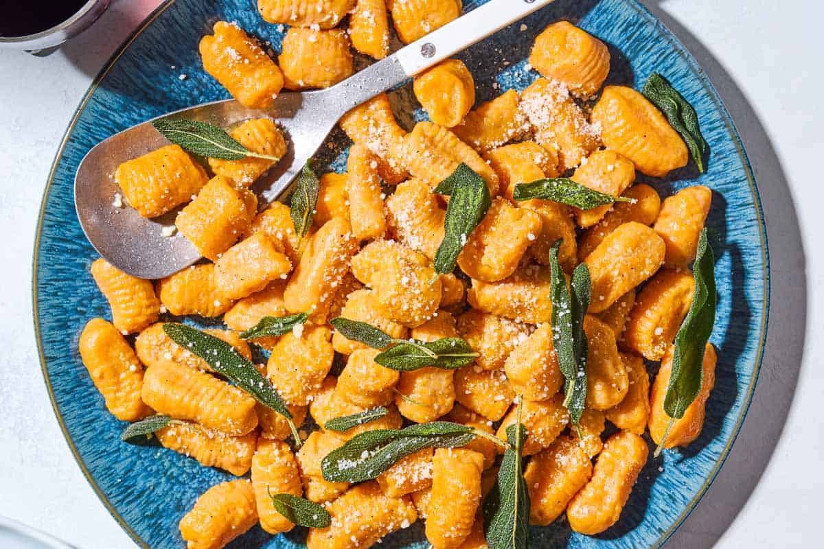 A close up of sweet potato gnocchi and sauce topped with fresh sage on a serving platter with a serving spoon spoon.