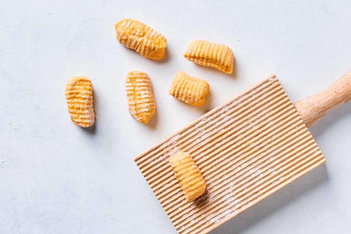 A sweet potato gnocchi being shaped on a wooden gnocchi board. Next to this is 4 more pieces of the gnocchi.