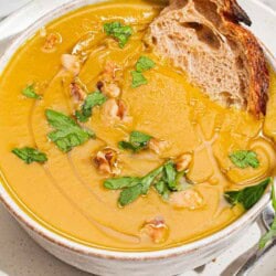 A close up of acorn squash soup garnished with walnuts, parsley and a drizzle of olive oil in a bowl with a piece of bread. Next to this is a spoon.