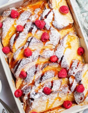 An overhead photo of baked french toast topped with powdered sugar and raspberries and drizzled with honey simple syrup in a baking dish.