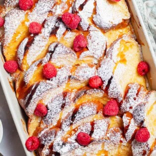 An overhead photo of baked french toast topped with powdered sugar and raspberries and drizzled with honey simple syrup in a baking dish.