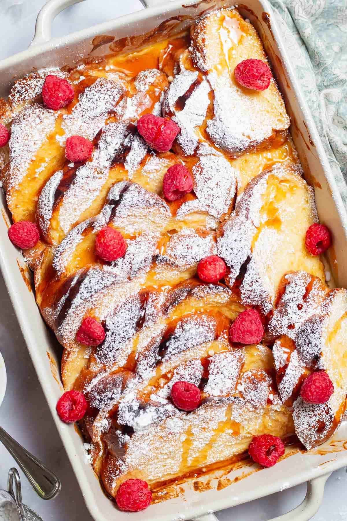 An overhead photo of baked french toast topped with powdered sugar and raspberries and drizzled with honey simple syrup in a baking dish.