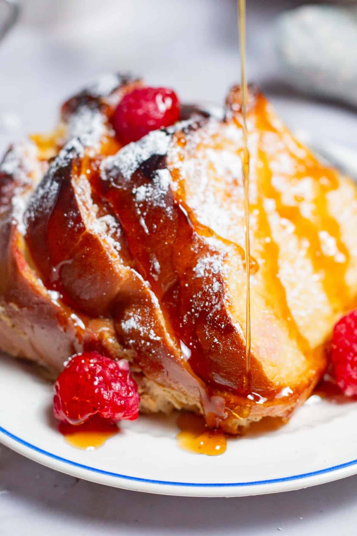 A close up of slices of baked french toast topped with raspberries and powdered sugar being drizzled with honey simple syrup on a plate.