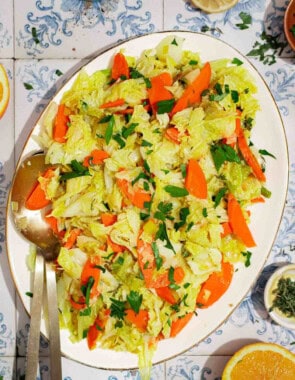 An overhead photo of braised savoy cabbage on a serving platter with serving utensils. Next to this is a orange wheel, bowls of black pepper and thyme, and a lemon half.