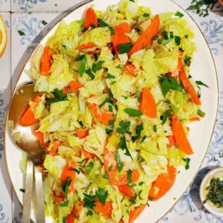 An overhead photo of braised savoy cabbage on a serving platter with serving utensils. Next to this is a orange wheel, bowls of black pepper and thyme, and a lemon half.