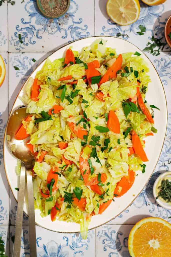 An overhead photo of braised savoy cabbage on a serving platter with serving utensils. Next to this is a orange wheel, bowls of black pepper and thyme, and a lemon half.