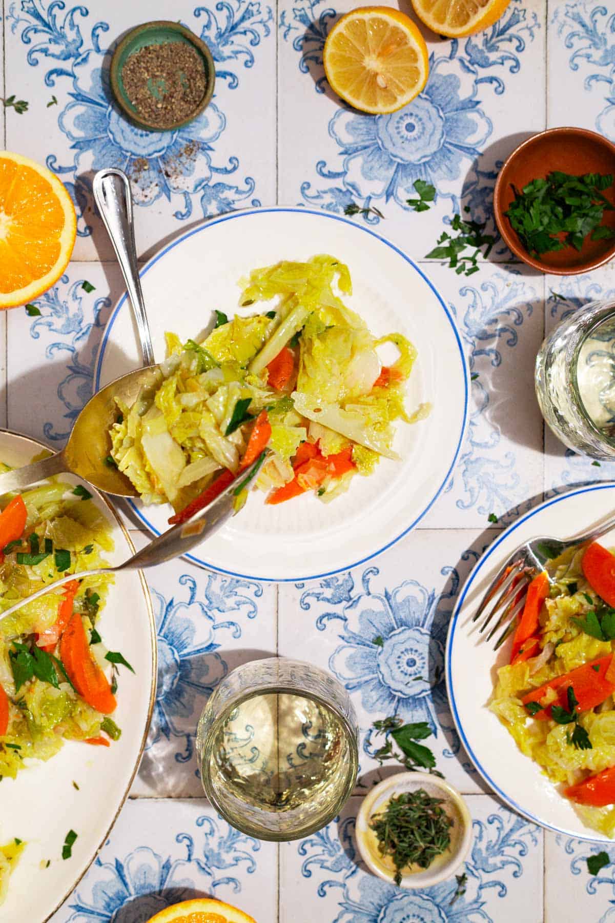 An overhead photo of a serving of braised savoy cabbage being placed a plate with a fork. Next to this is another plate of the cabbage, a platter of the cabbage, a glass of wine, bowls of black pepper, parsley and thyme, an orange wheel, and 2 lemon halves.