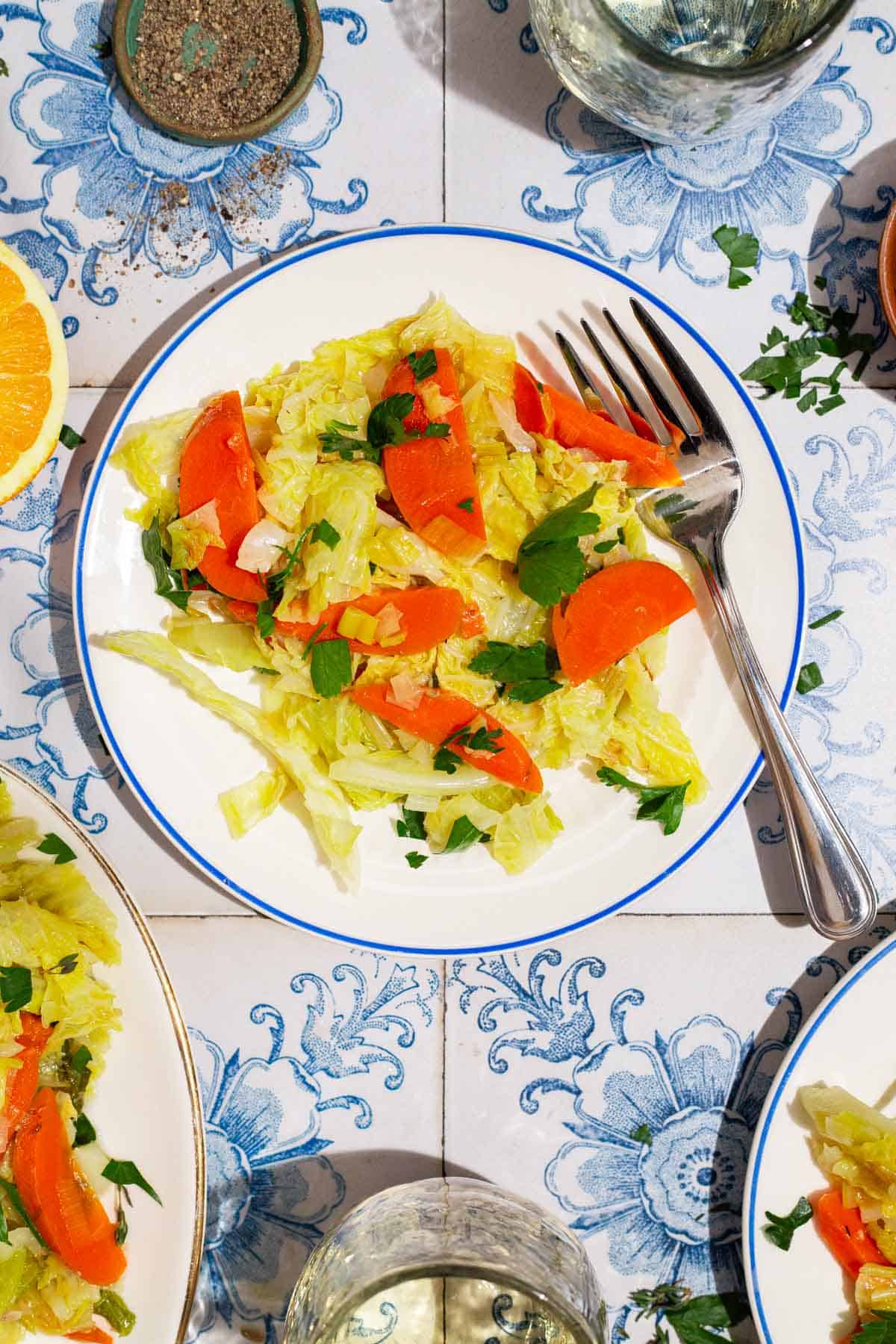 An overhead photo of a serving of braised savoy cabbage on a plate with a fork. Next to this is a bowl of black pepper and 2 glasses of wine.