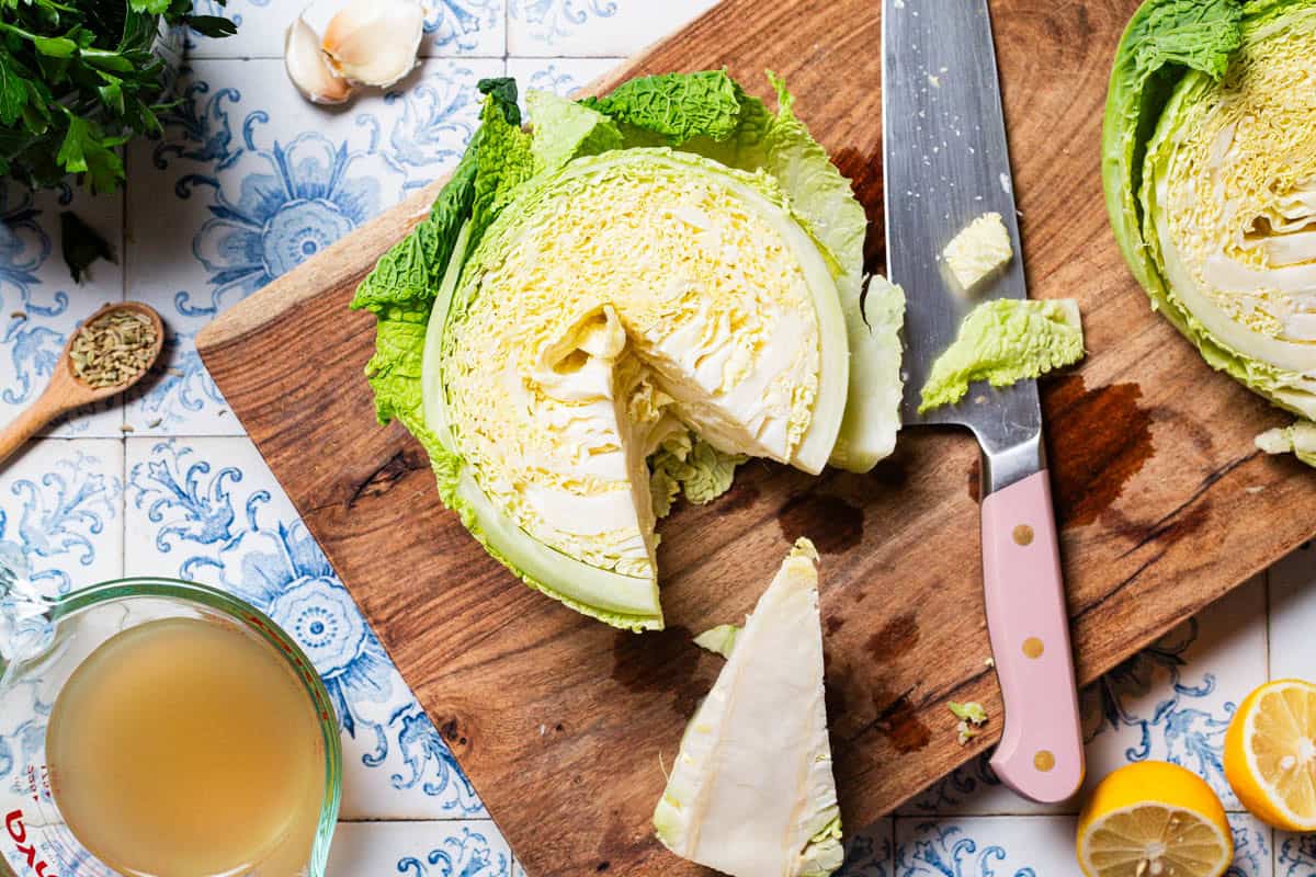 An overhead photo of a half a head of savoy cabbage with the core removed on a cutting board. Next to this is a knife, the other half of a cabbage, 2 lemon halves, garlic cloves, parsley, a spoon with fennel seeds, and a measuring cup with broth.