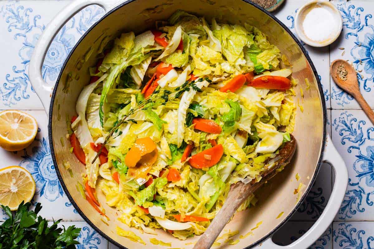 Braised savoy cabbage in a pot with a wooden spoon. Next to this is a bowl of salt, 2 lemon halves, a wooden spoon with fennel seeds, and some parsley.