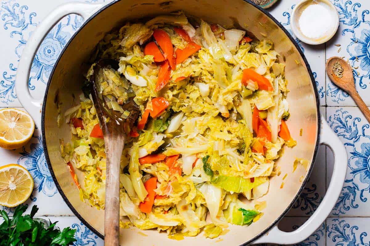 Braised savoy cabbage in a pot with a wooden spoon. Next to this is a bowl of salt, 2 lemon halves, a wooden spoon with fennel seeds, and some parsley.