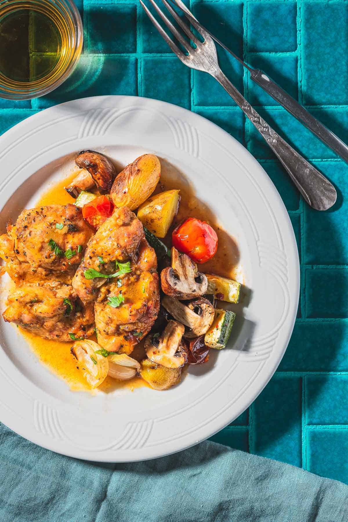 A serving of garlic dijon chicken with roasted vegetable on a plate. Next to this is a cloth napkin, fork, knife and a glass of wine.