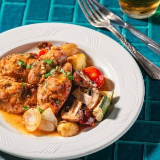 A serving of garlic dijon chicken with roasted vegetable on a plate. Next to this is a cloth napkin, fork, knife and a glass of wine.