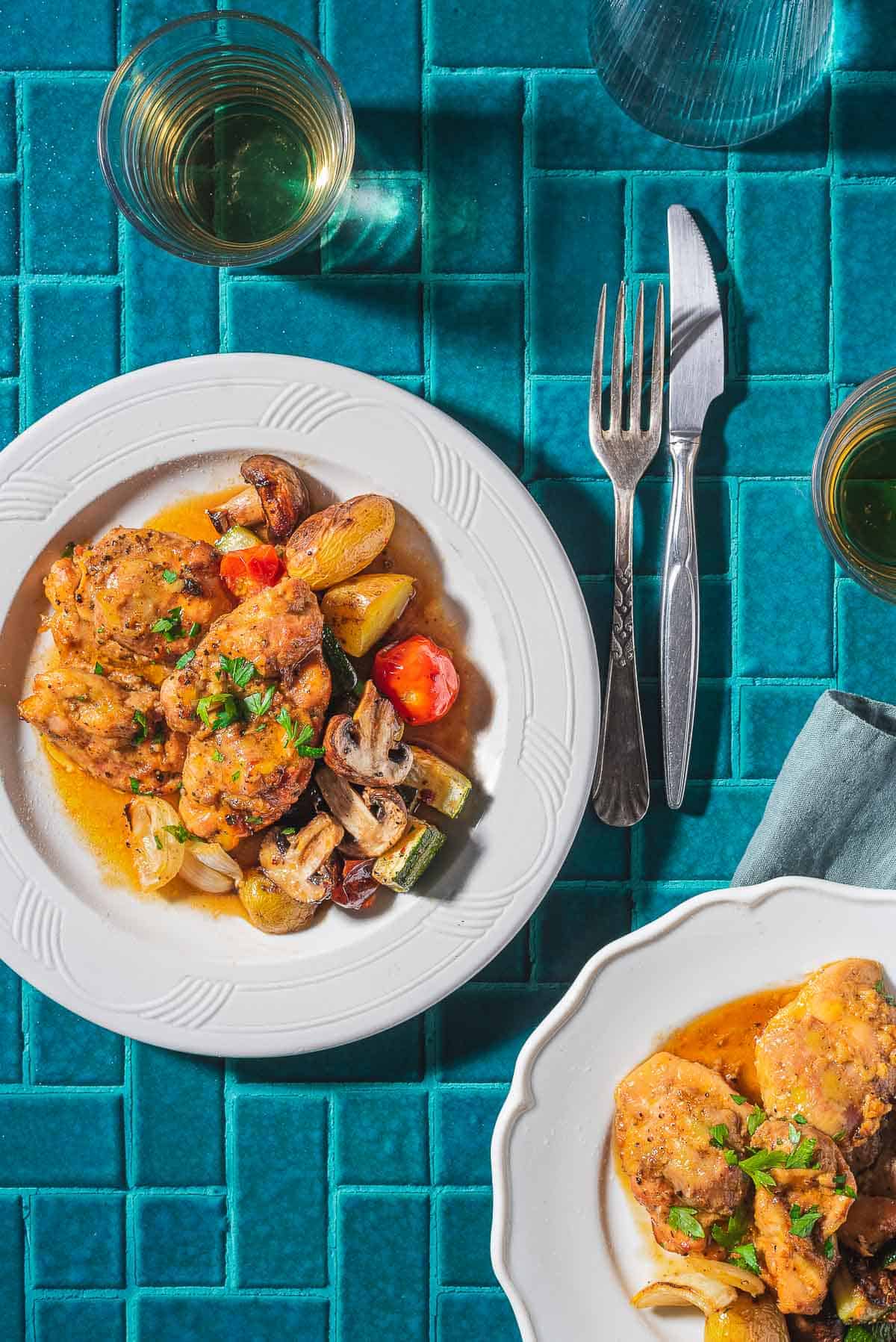 2 servings of garlic dijon chicken with roasted vegetables on plates. Next to these are 2 glasses of wine, a cloth napkin, fork and knife.