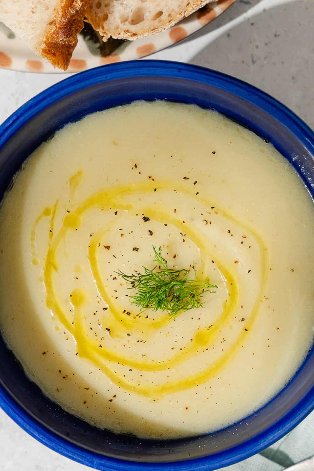 An overhead photo of a bowl of fennel soup topped with a drizzle of olive oil and fennel fronds. Next to this is a plate of sliced bread.