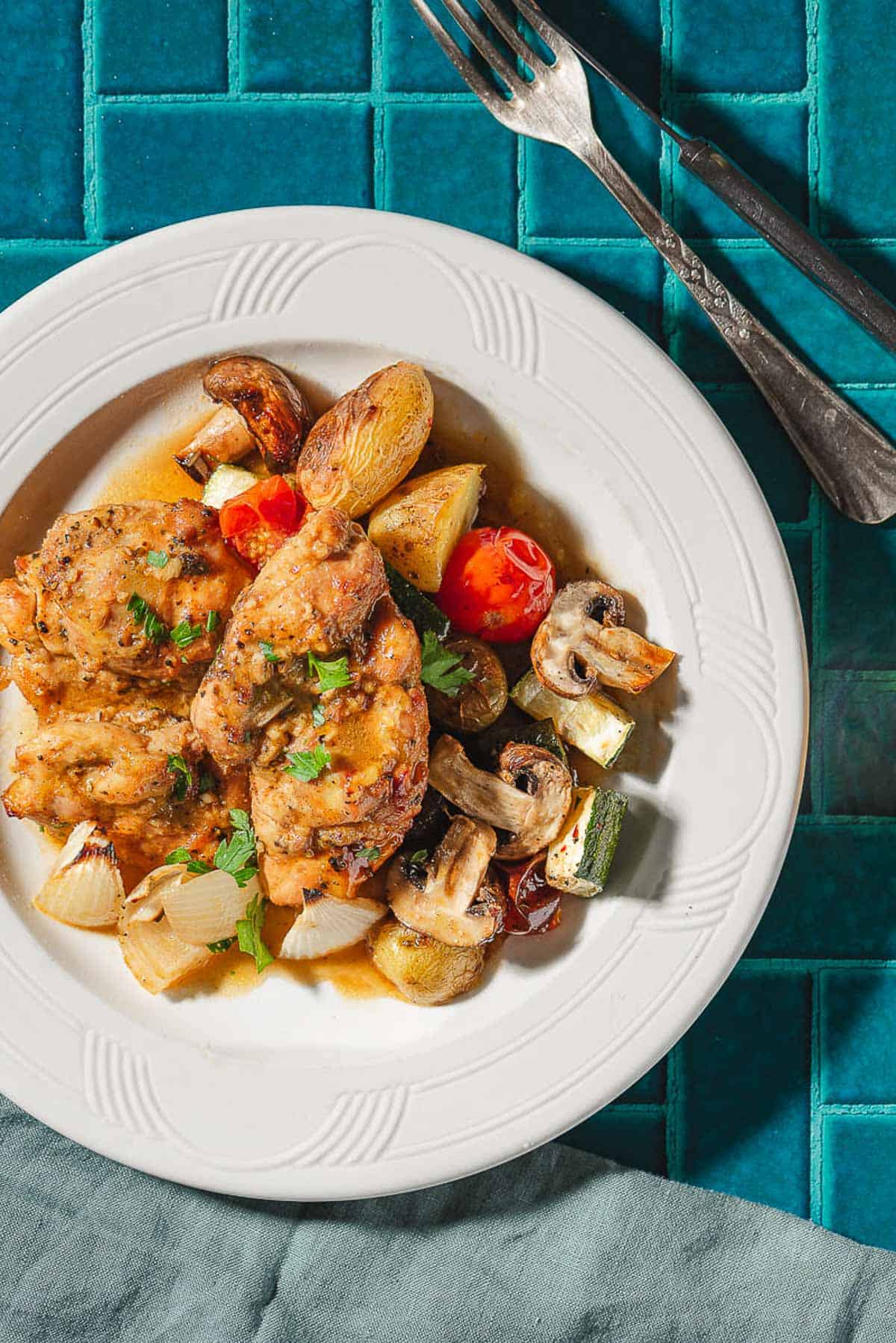 A serving of garlic dijon chicken with roasted vegetable on a plate. Next to this is a cloth napkin, fork and knife.