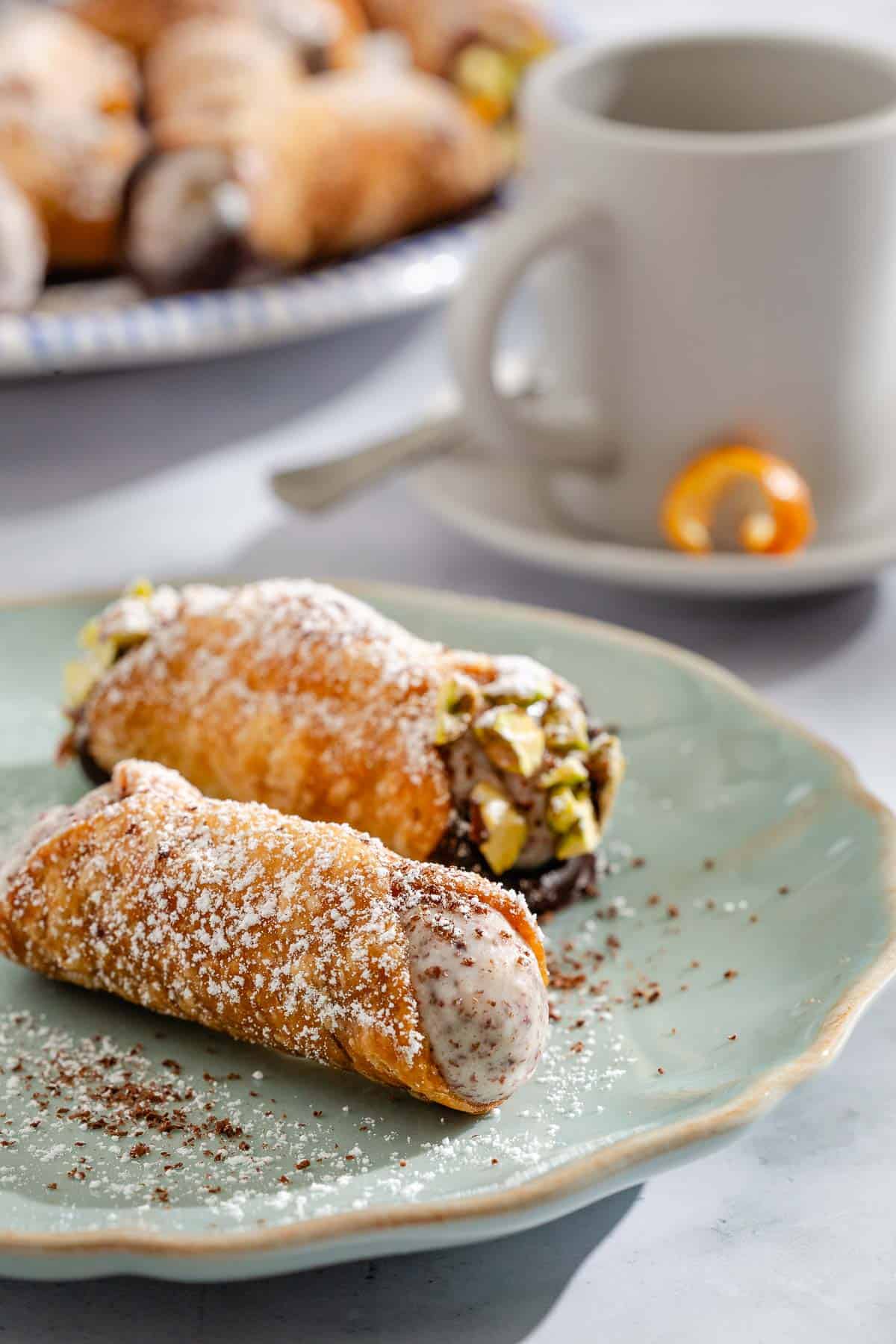 A close up of 2 cannolis on a plate. In the background is a mug on a plate with a spoon and a plate with the rest of the cannolis.