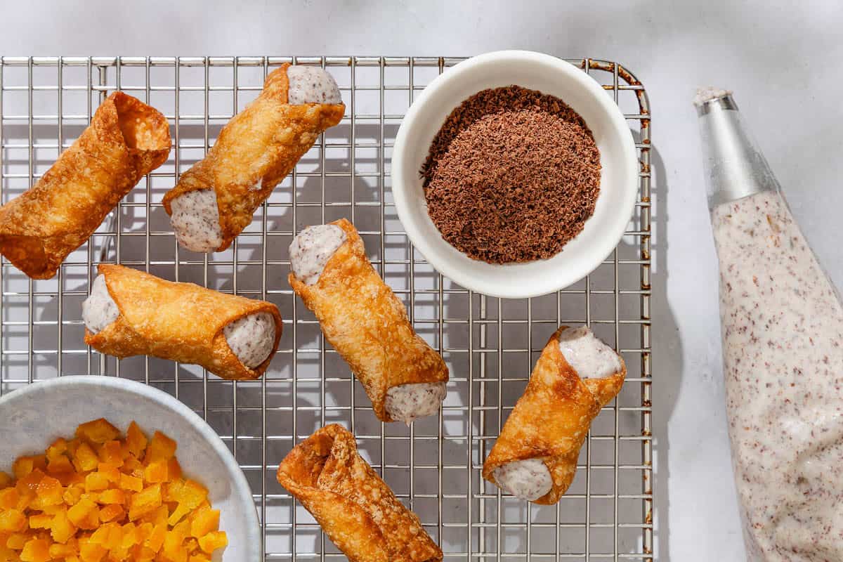 Several filled cannoli and some cannoli shells on a wire rack with bowls of grated dark chocolate and candied fruit.