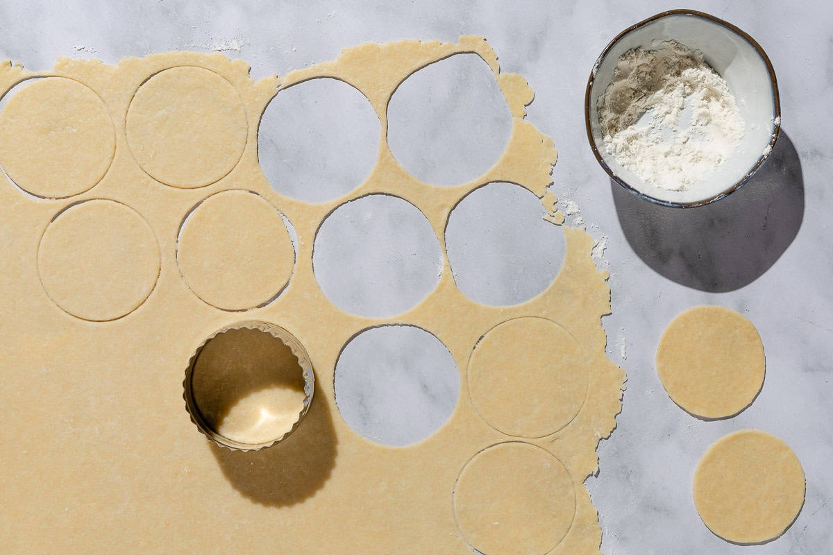 An overhead photo of pastry circles being cut out of rolled dough. Next to this is a bowl of flour.