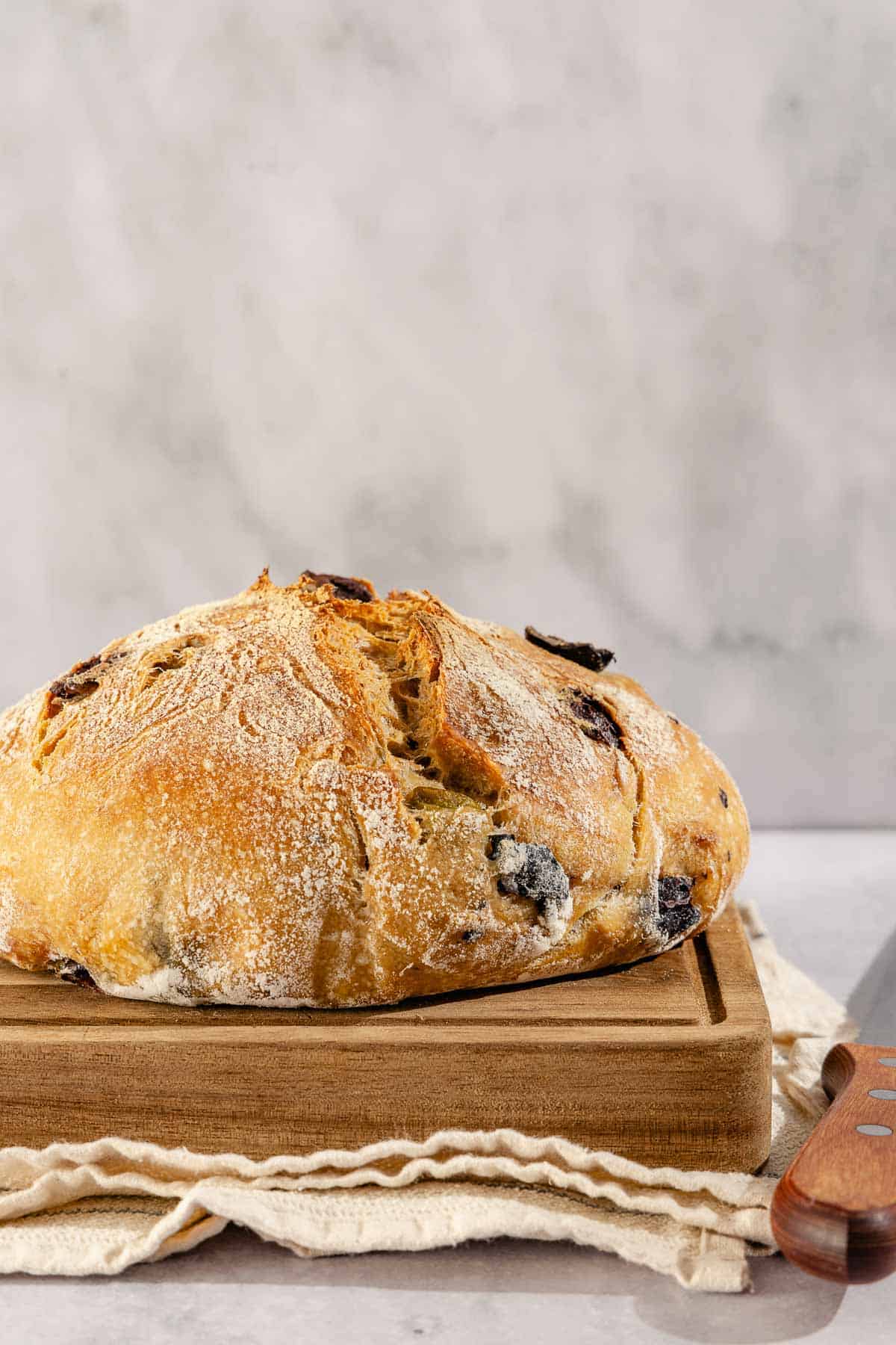 A close up photo of a loaf of olive bread on a cutting board sitting on a kitchen towel next to a knife.