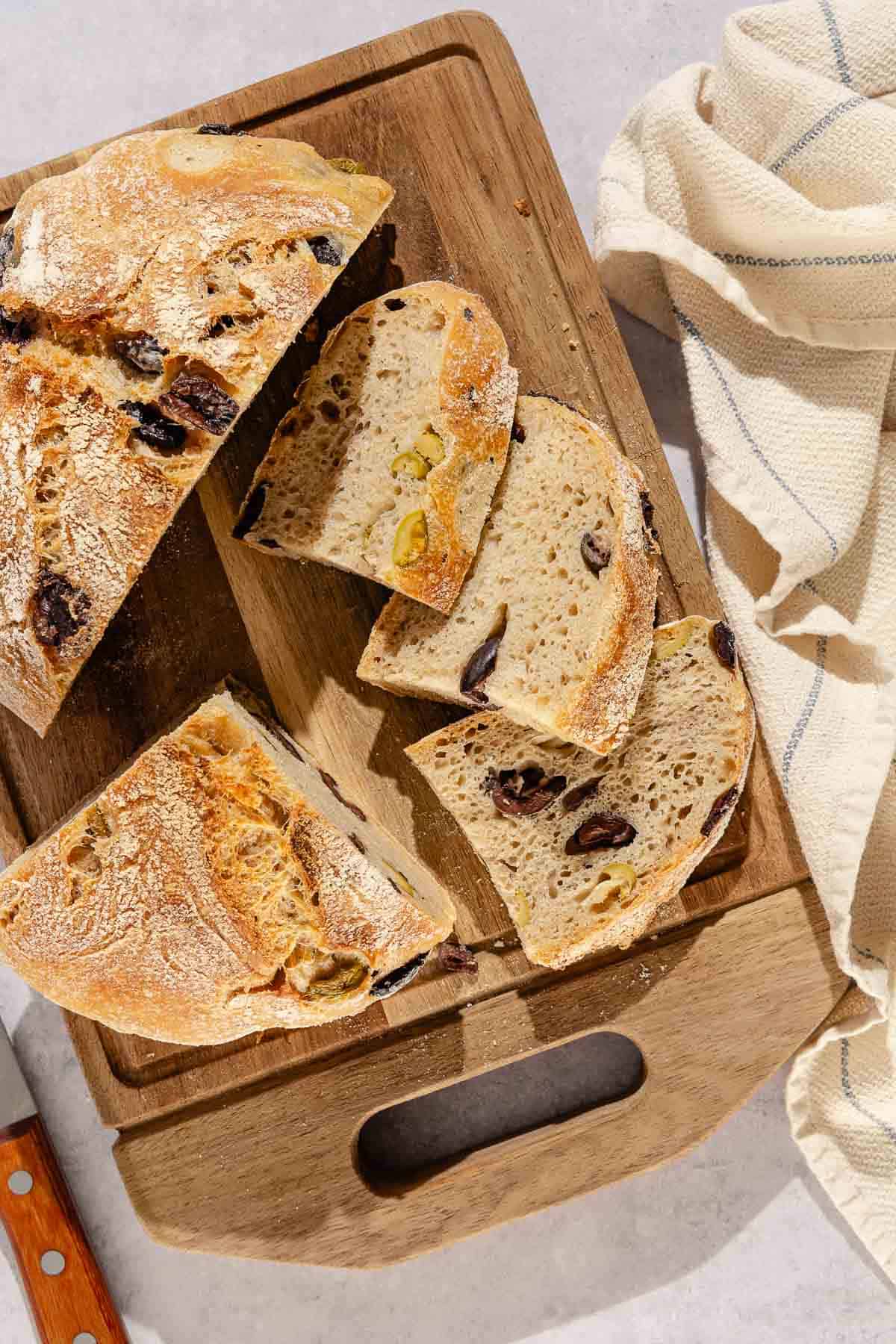 An overhead photo of half a loaf of olive bread next to slices of olive bread on a wooden cutting board. Next to this is a knife and a kitchen towel.