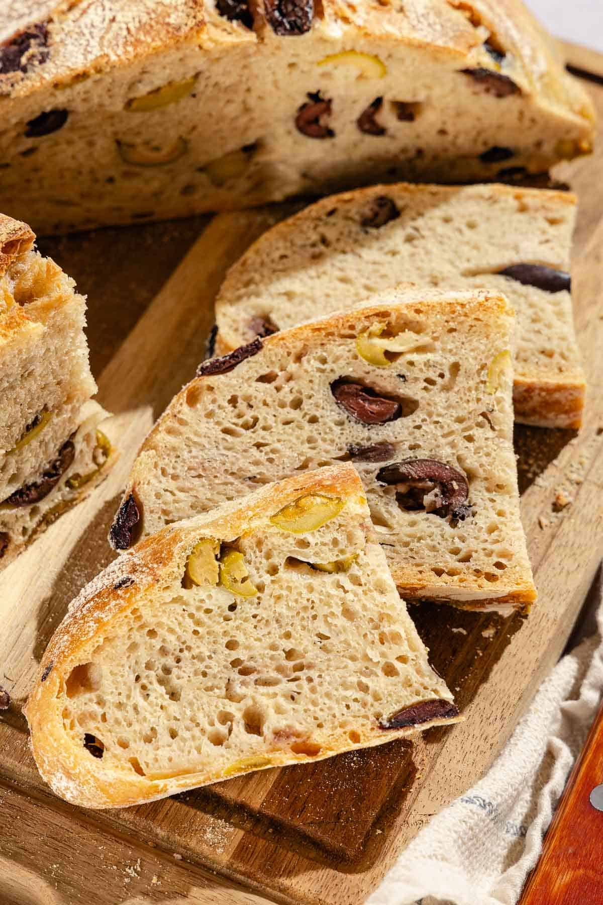 A close up photo of 3 slices of olive bread and half of a loaf on a wooden cutting board.