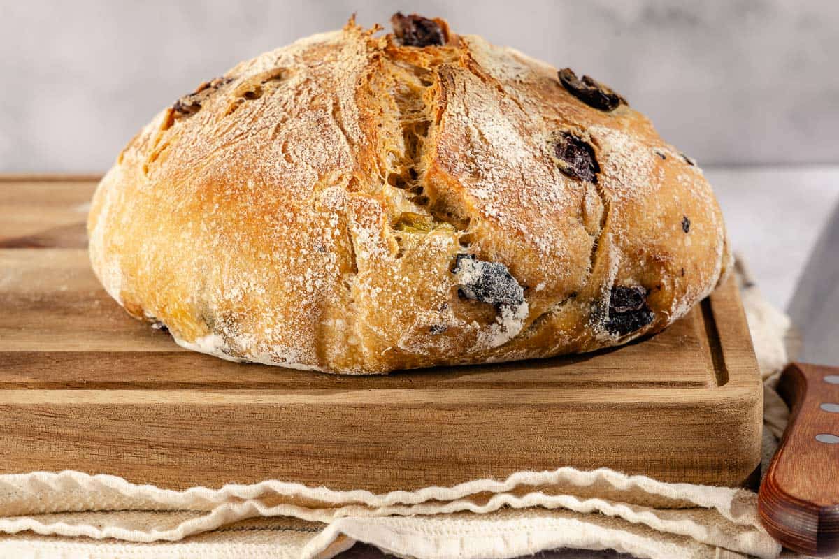 A close up photo of a loaf of olive bread on a cutting board sitting on a kitchen towel next to a knife.