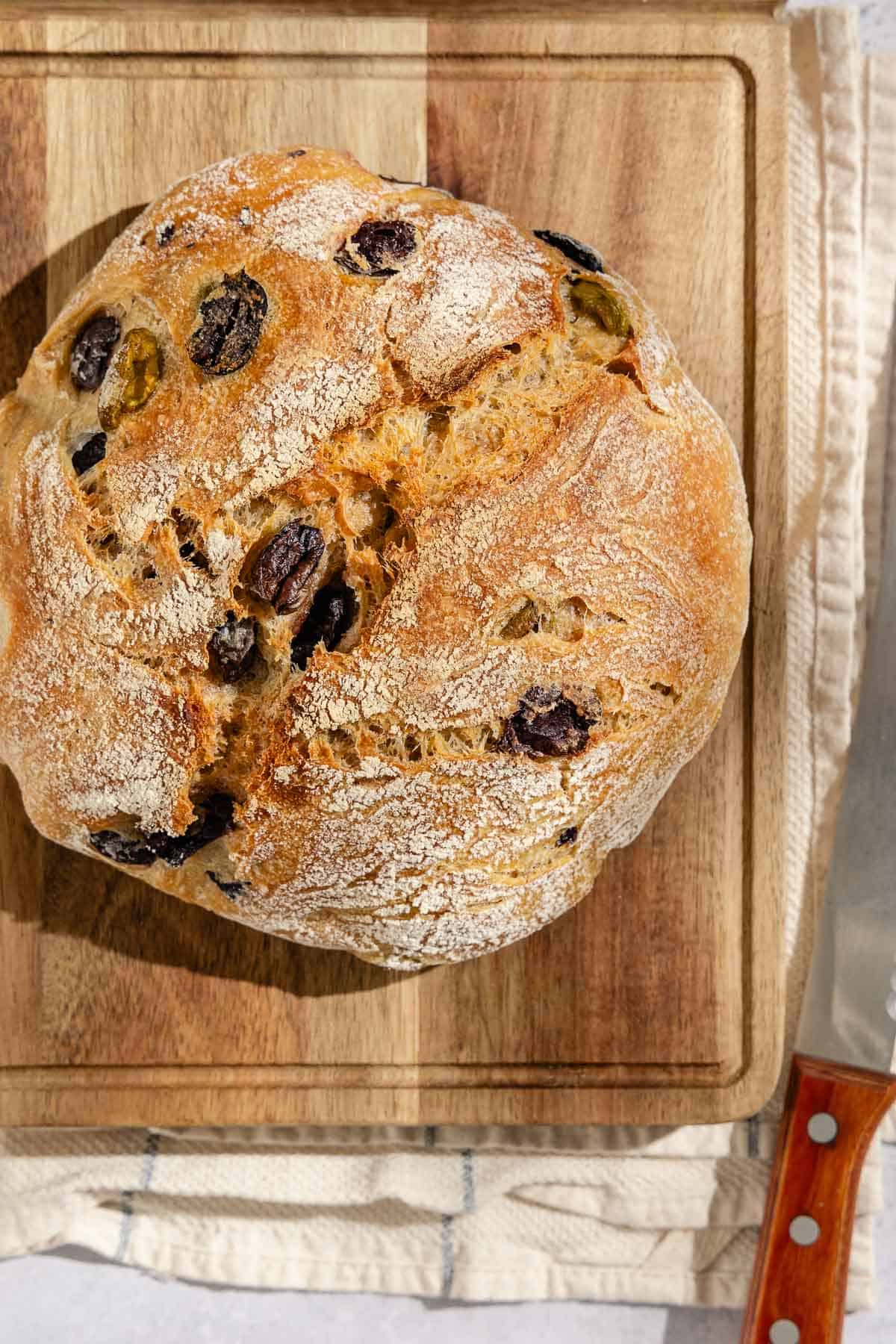 An overhead photo of a loaf of olive bread on a cutting board sitting on a kitchen towel next to a knife.