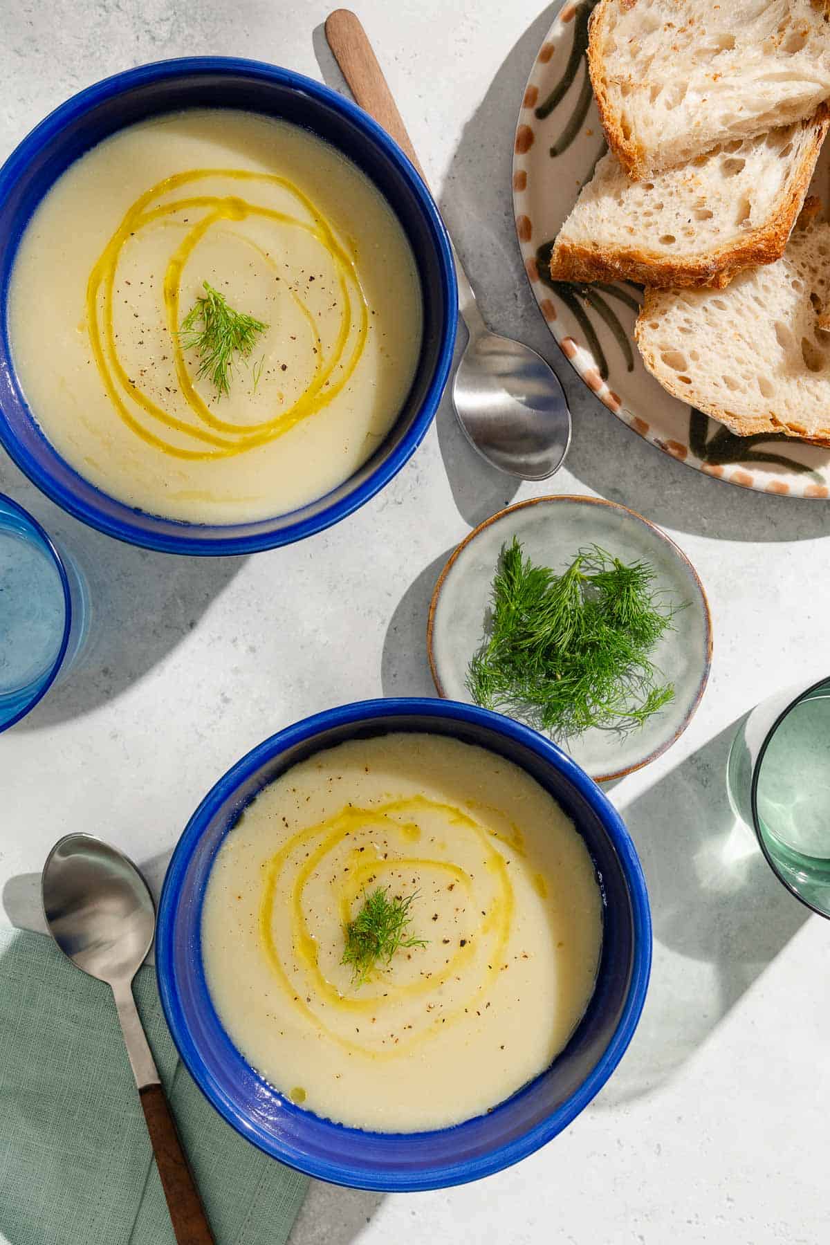 An overhead photo of 2 bowls of fennel soup topped with a drizzle of olive oil and fennel fronds. Next to these are spoons and a cloth napkin, plates of sliced bread and fennel fronds, and 2 glasses of water.