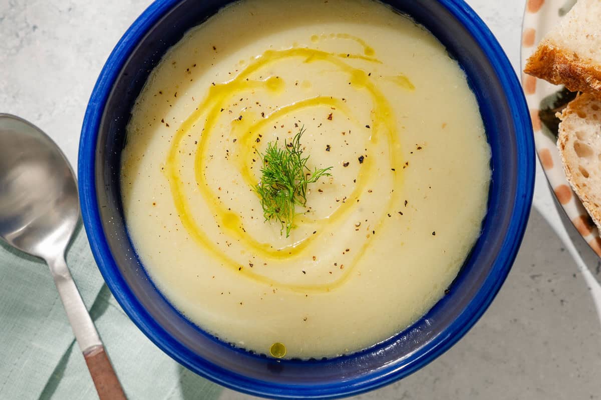 An overhead photo of a bowl of fennel soup topped with a drizzle of olive oil and fennel fronds. Next to this is a cloth napkin, spoon and a plate of sliced bread.