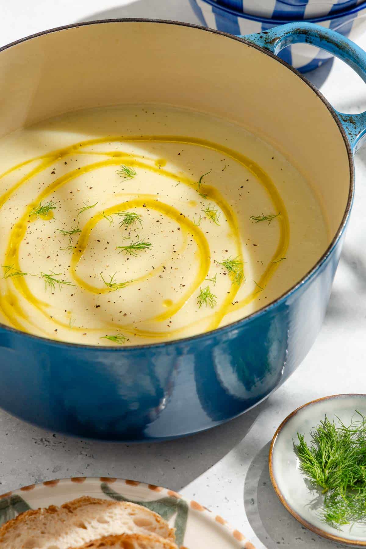 A pot of fennel soup topped with a drizzle of olive oil and fennel fronds. Next to this are plates with sliced bread and fennel fronds.