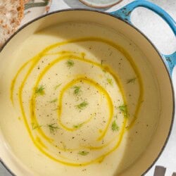 An overhead photo of a pot of fennel soup topped with a drizzle of olive oil and fennel fronds. Next to this are plates of sliced bread and fennel fronds, as well as an empty bowl holding 2 spoons.