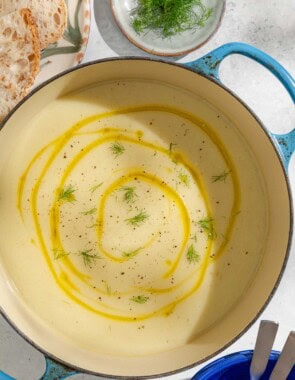 An overhead photo of a pot of fennel soup topped with a drizzle of olive oil and fennel fronds. Next to this are plates of sliced bread and fennel fronds, as well as an empty bowl holding 2 spoons.