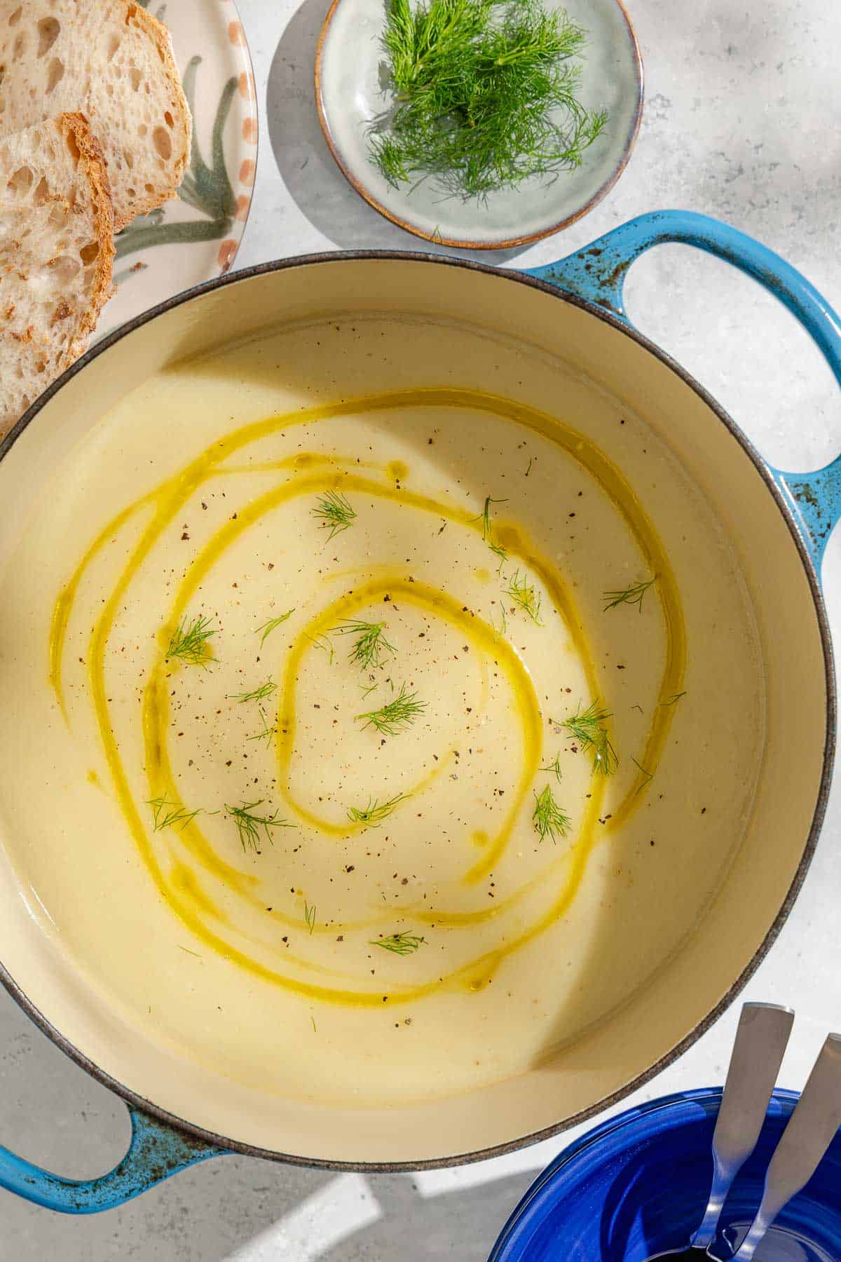 An overhead photo of a pot of fennel soup topped with a drizzle of olive oil and fennel fronds. Next to this are plates of sliced bread and fennel fronds, as well as an empty bowl holding 2 spoons.