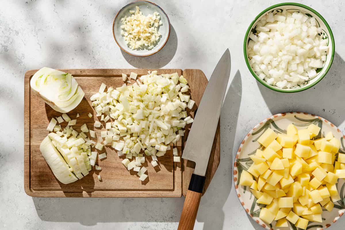 An overhead photo of fennel bulbs and diced fennel on a cutting board with a knife. Next to this are bowls of chopped potatoes, onions and garlic.