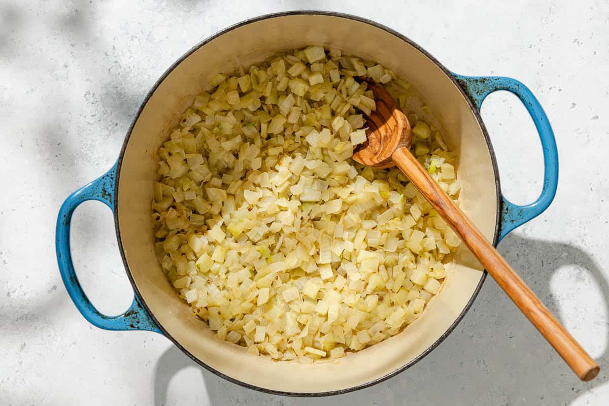 An overhead photo of diced onions and fennel being sauteed in a large pot with a wooden spoon.