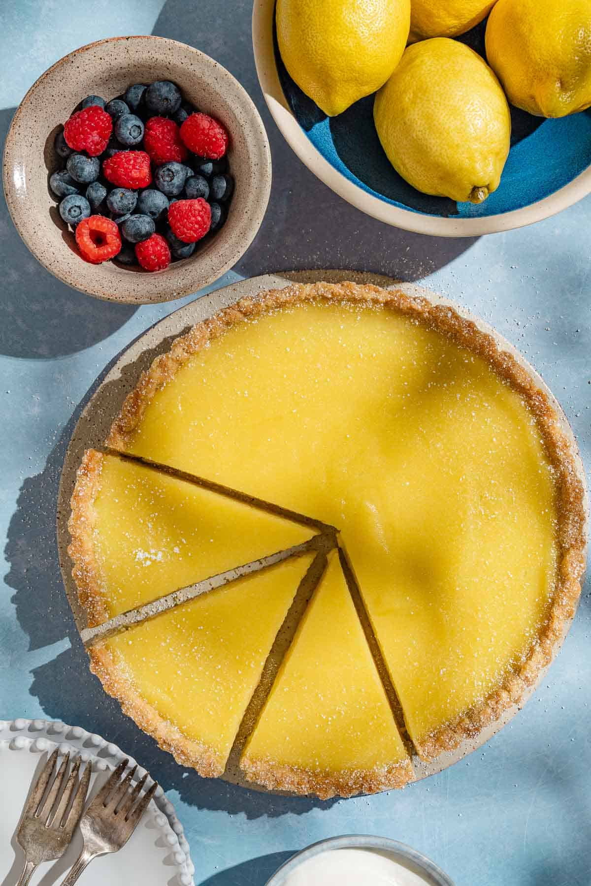 An overhead photo of a lemon tart with 3 pieces sliced. Next to this is a stack of 2 plates with 2 forks, and bowls of lemons, and berries.