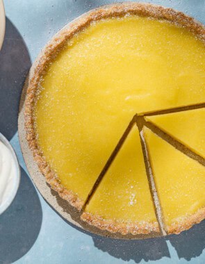 An overhead photo of a lemon tart with 3 pieces sliced. Next to this is a stack of 3 plates with 2 forks, and bowls of lemons, berries and greek yogurt.