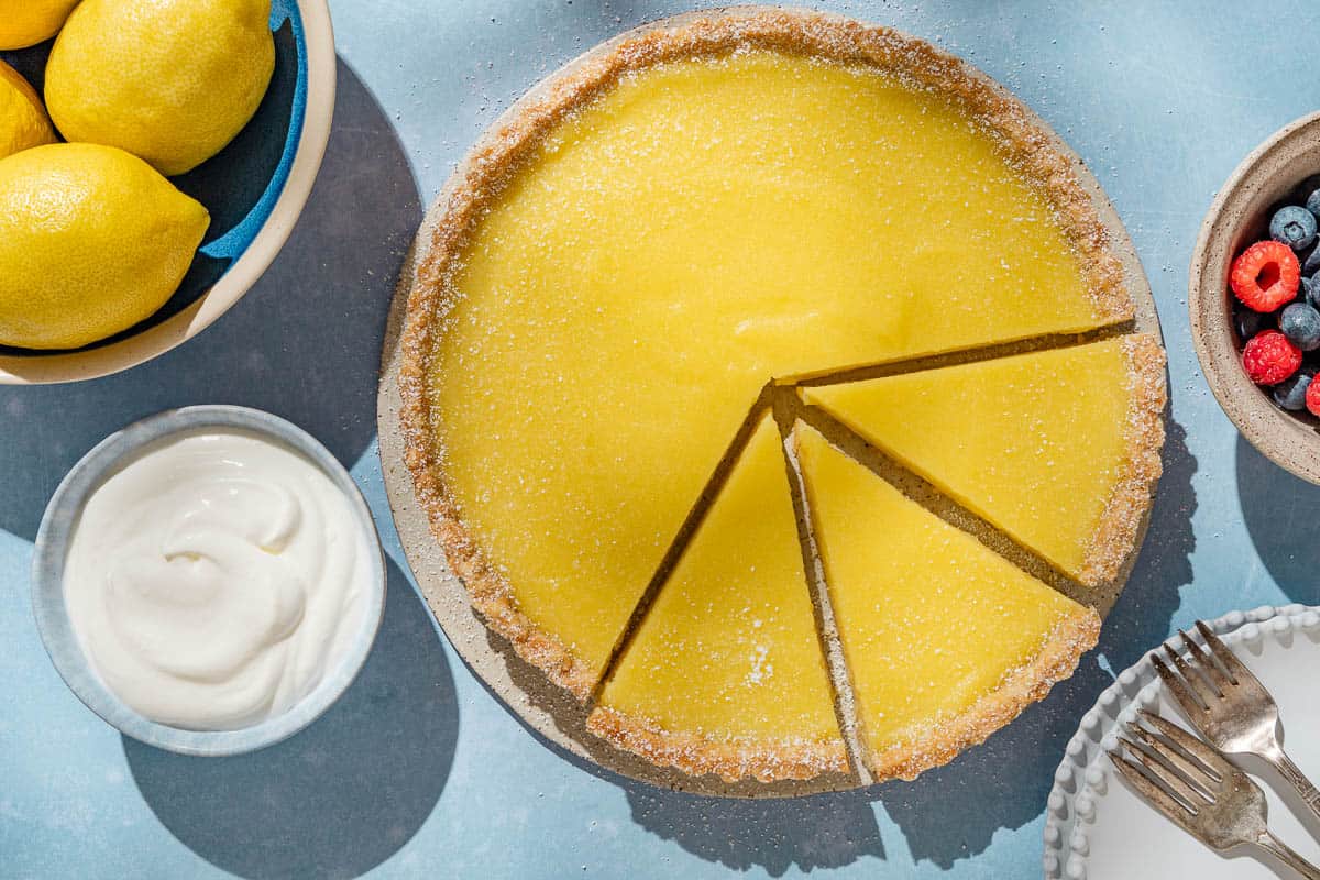 An overhead photo of a lemon tart with 3 pieces sliced. Next to this is a stack of 3 plates with 2 forks, and bowls of lemons, berries and greek yogurt.