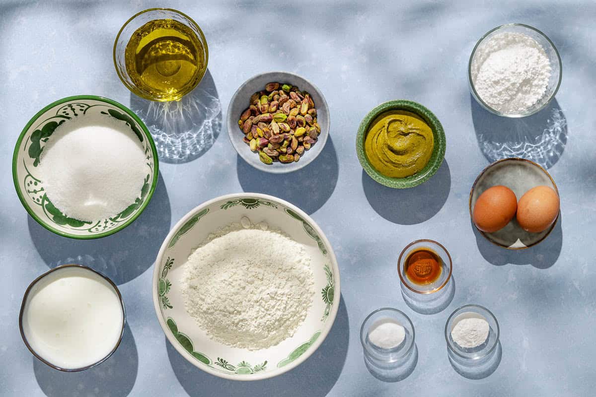 Ingredients for pistachio cake including pistachios, flour, baking powder, baking soda, salt, greek yogurt, granulated sugar, eggs, olive oil, vanilla extract, pistachio butter, and powdered sugar.