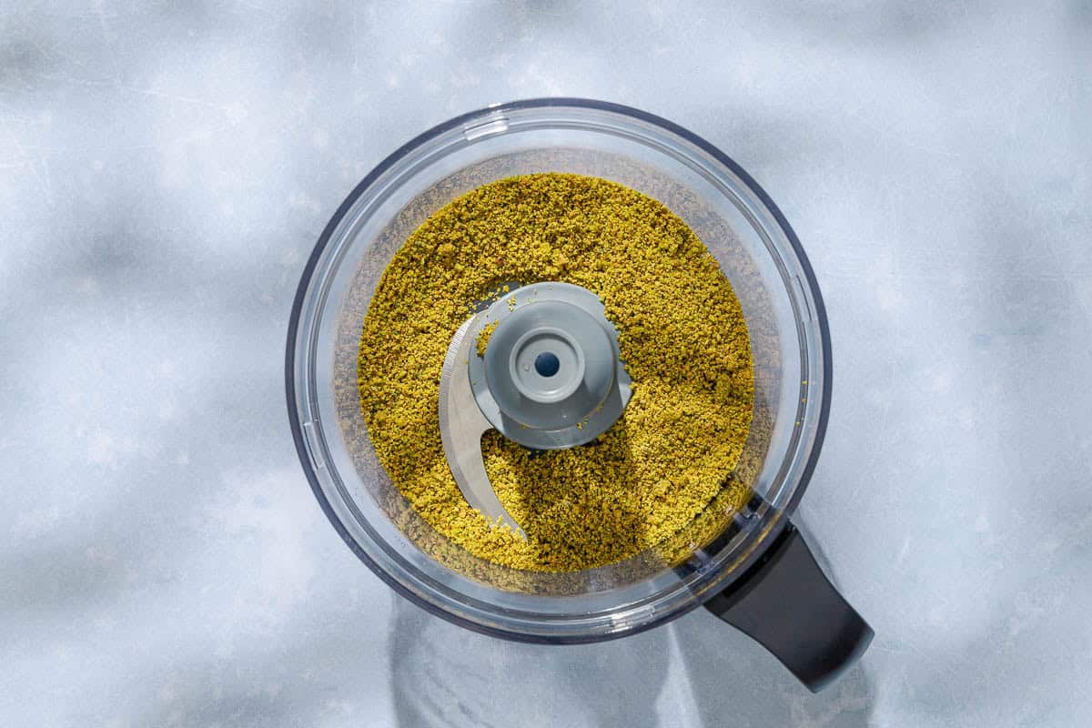 An overhead photo of ground pistachios in the bowl of a food processor fitted with a blade.