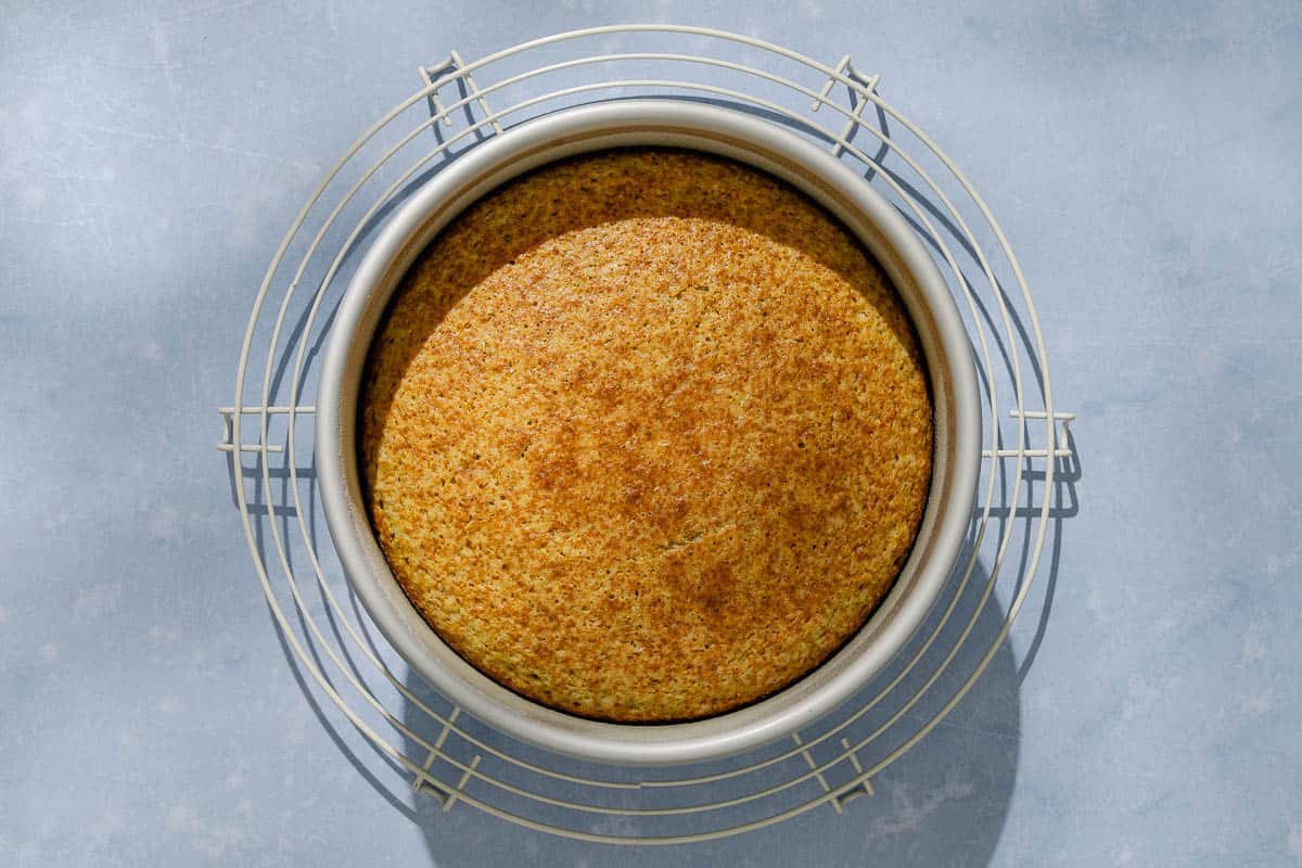 An overhead photo of the unglazed, baked pistachio cake in a round baking pan cooling on a wire rack.