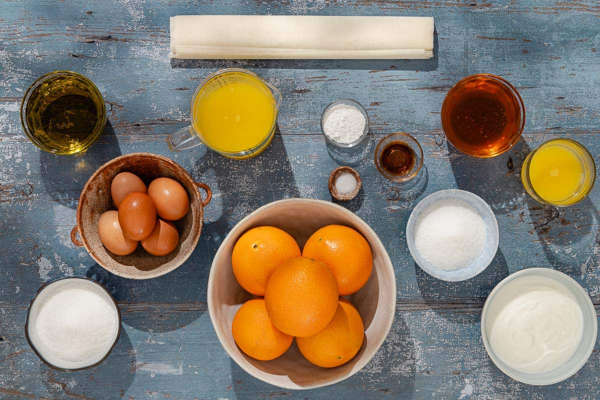 Ingredients for portokalopita greek orange cake including orange juice, honey, sugar, phyllo, eggs, baking powder, olive oil, plain yogurt, oranges, vanilla extract and salt.