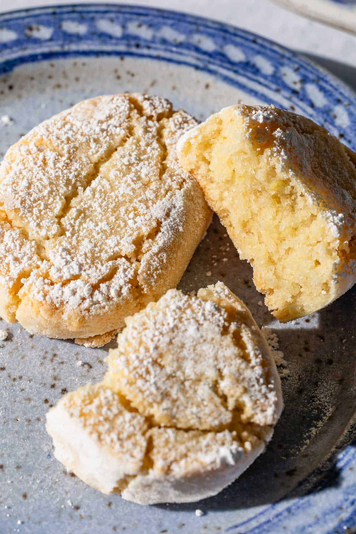 A close up of 2 baked ricciarelli cookies topped with powdered sugar on a plate. One is split in half.